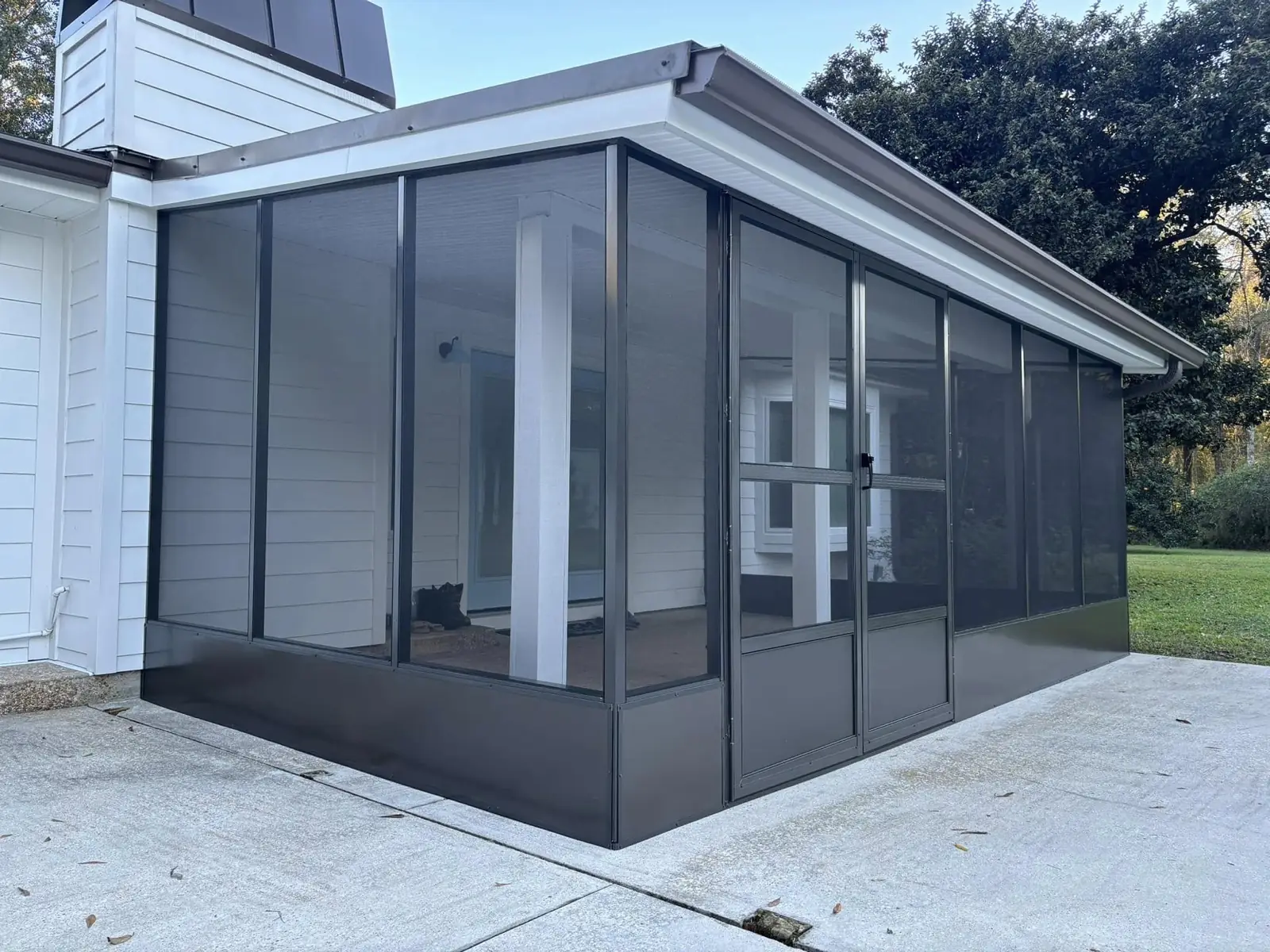 Bronze-frame glass room with a hip roof attached to the side of a white-siding home over a concrete slab, built by Tiger Town Construction